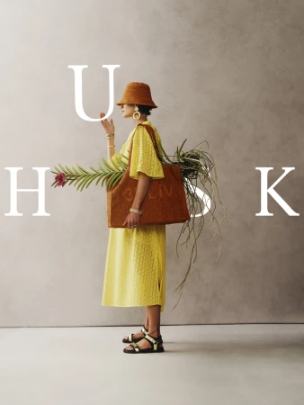 A stylish adult female is posing with a brown tote bag, wearing a yellow dress, straw hat, and sandals against a neutral backdrop.