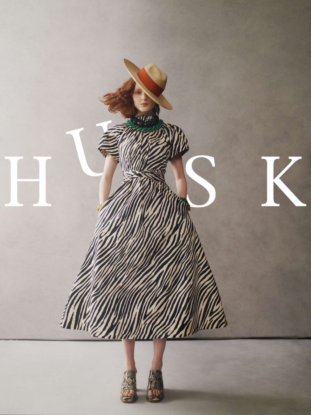A red-haired woman stands in a zebra-striped dress and hat against a grey backdrop, featuring the brand 'HUSK' in large white letters.
