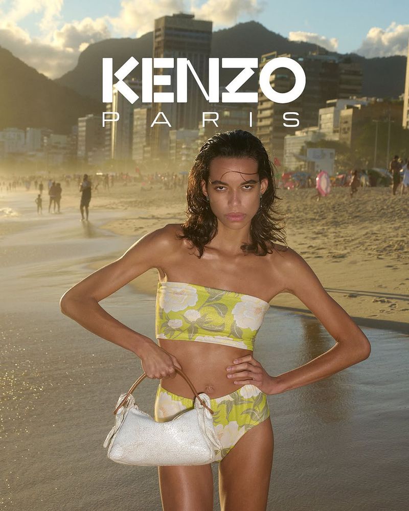 Woman in floral bikini holds purse on sandy beach with city skyline backdrop, posing in the sunlight. Kenzo Paris logo is visible.