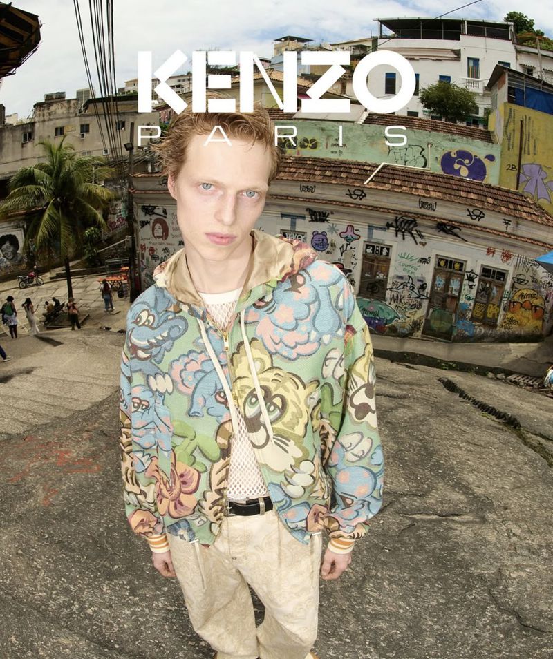 Male fashion model in a patterned hooded jacket and tan pants poses on a stone street in Rio, with hillside buildings.