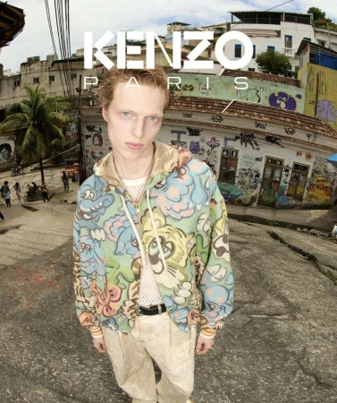 Male fashion model in a patterned hooded jacket and tan pants poses on a stone street in Rio, with hillside buildings.