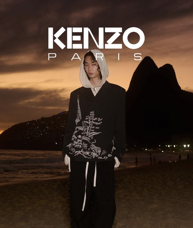 Fashionable person in KENZO jacket standing on beach with mountain backdrop at dusk, styled for a high-end editorial campaign.