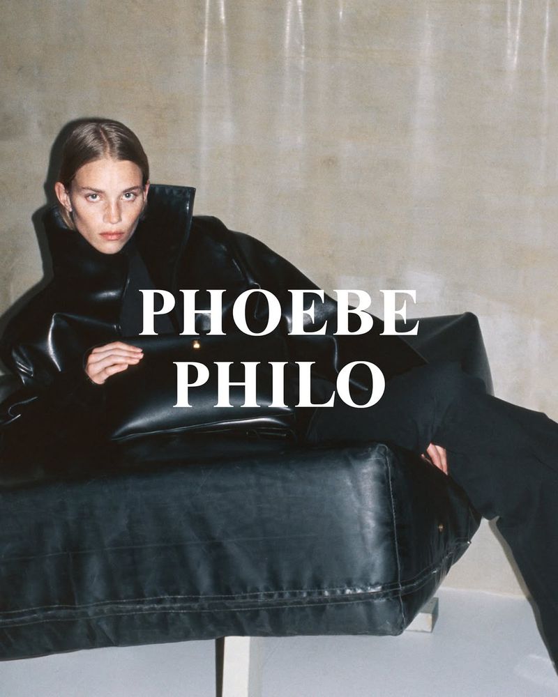 Fashion shot of a model in a black leather jacket reclining on a black leather couch against a neutral backdrop, advertising Phoebe Philo.
