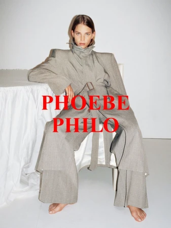 Fashionable adult female poses in a gray belted outfit and bare feet against a light background while seated with hands resting on a table.