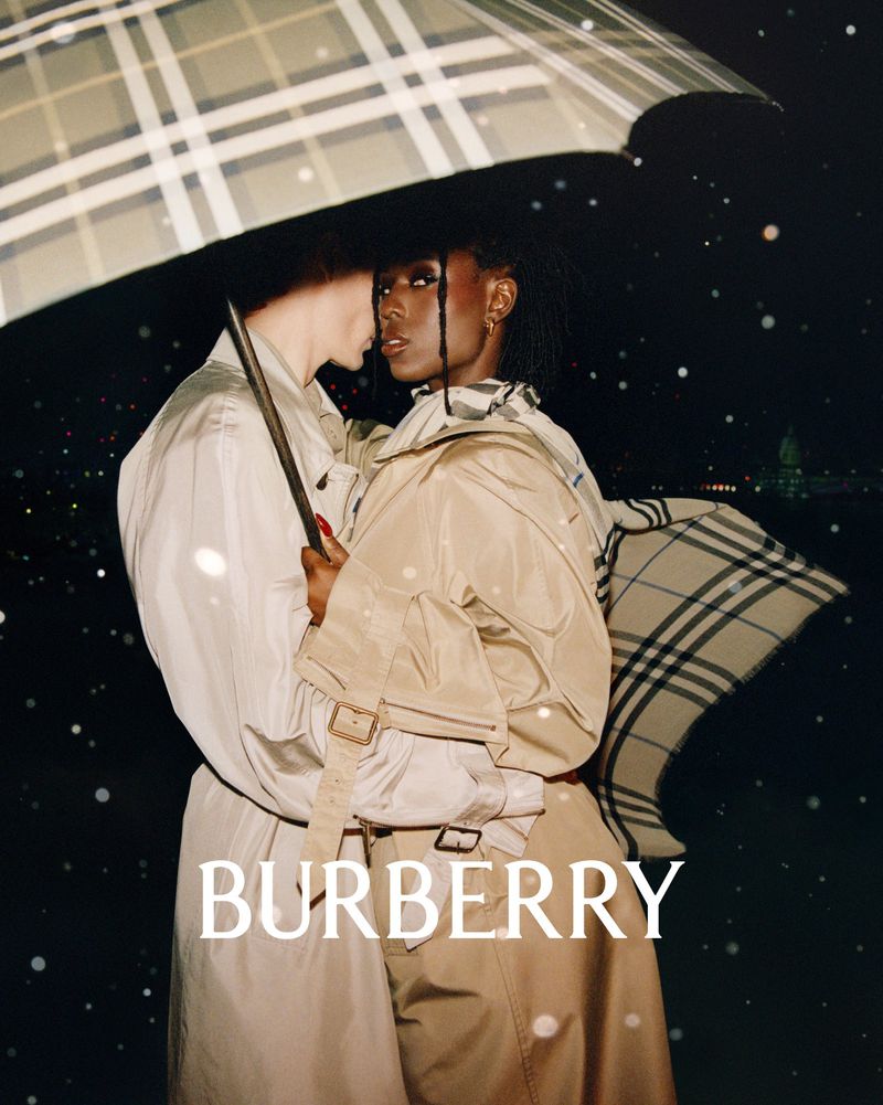 Two people standing closely under a checkered umbrella at night wearing beige trench coats and scarves for Burberry London in Love