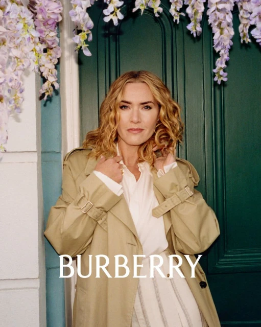 A fair-skinned woman with curly blonde hair, wearing a trench coat, poses in front of a green door with lilac flowers above.