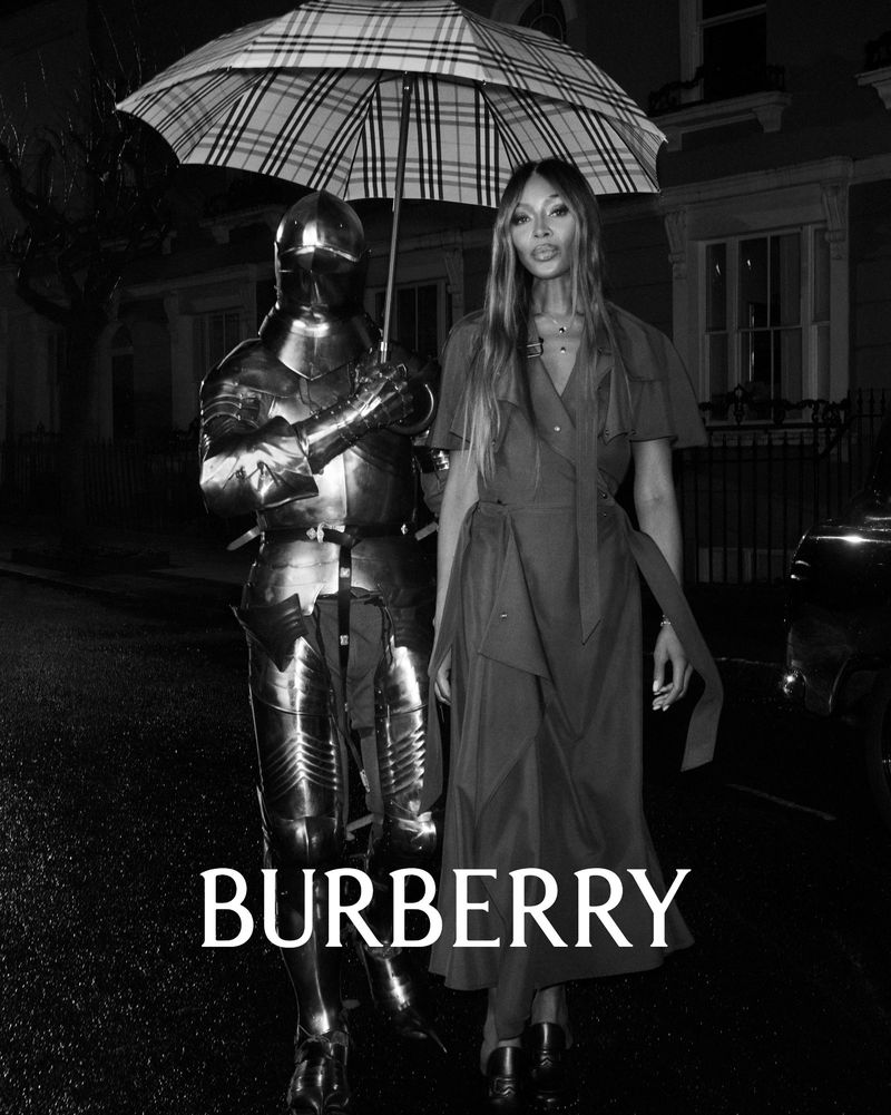 Naomi Campbell poses with a knight in full armor under an umbrella in an outdoor fashion shot, highlighting textures, shapes, and Burberry logo.