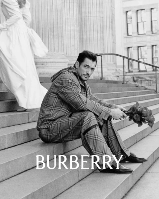 Black and white outdoor fashion shot of a male model in plaid suit sitting on stairs with roses, featuring a partial dress.
