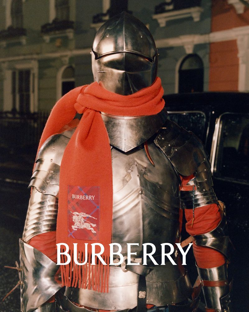 Person in silver armour with red scarf stands in front of a car on a street with residential buildings in the background.