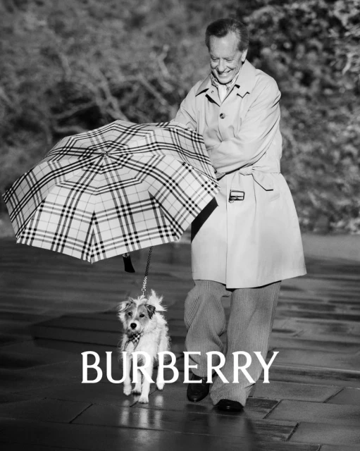 A person in a tan trench coat smiles while walking a dog under a Burberry umbrella on a city street, black and white fashion.