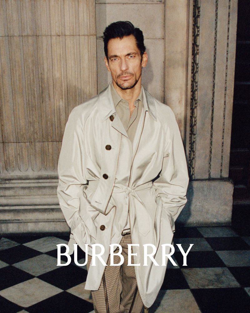 Fashion shot of a handsome male adult wearing a trench coat, standing in front of a column and a black and white tiled floor indoors.