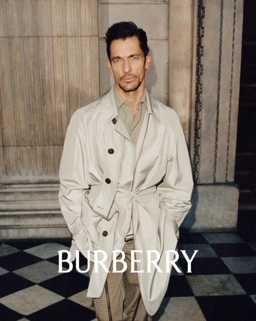 Fashion shot of a handsome male adult wearing a trench coat, standing in front of a column and a black and white tiled floor indoors.