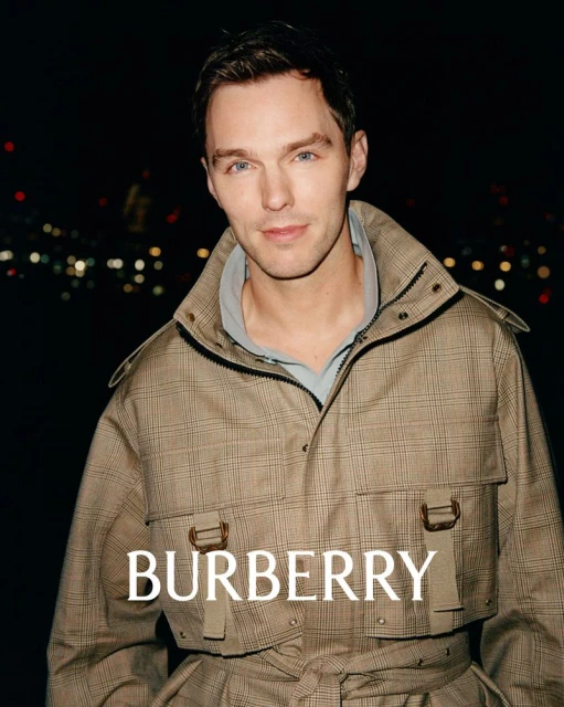 Nicholas Hoult poses in a Burberry trench coat against a bokeh-lit nightscape, fashion-forward and modern, capturing the brand's essence.