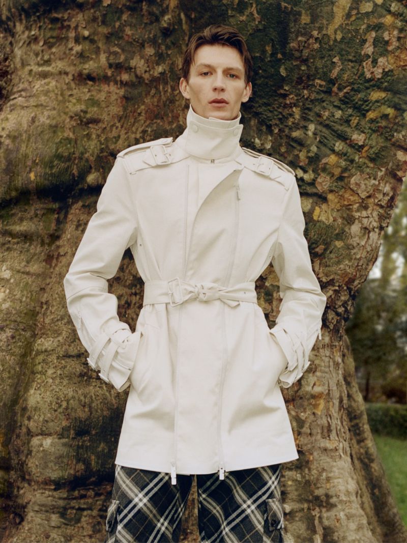 Fashion portrait of a slender adult male in a white jacket and plaid pants, posing against a large tree in a nature setting.