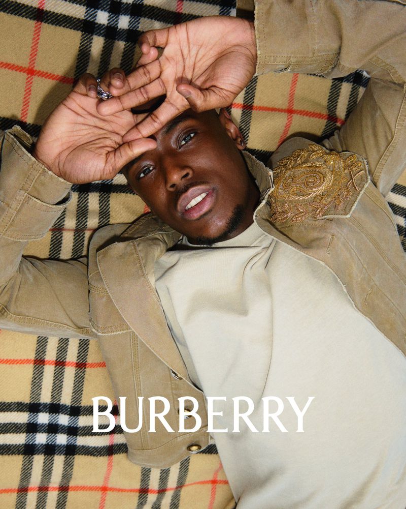 Black man lying down wearing a beige jacket and cream top against a classic plaid blanket background with Burberry text at the bottom.