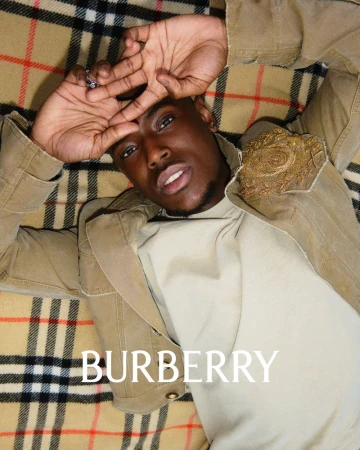 Black man lying down wearing a beige jacket and cream top against a classic plaid blanket background with Burberry text at the bottom.