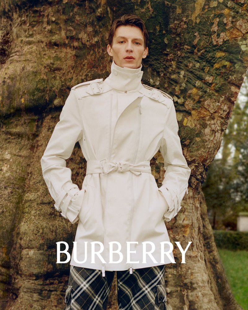 A young adult male model in a white trench coat poses in front of a large tree in a Burberry fashion campaign image.