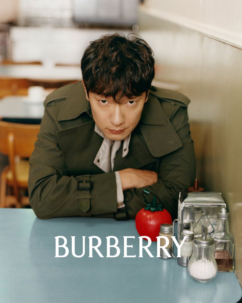 An Asian male model wearing a trench coat poses indoors at a table with salt and pepper shakers and a red apple. Fashion advertising.
