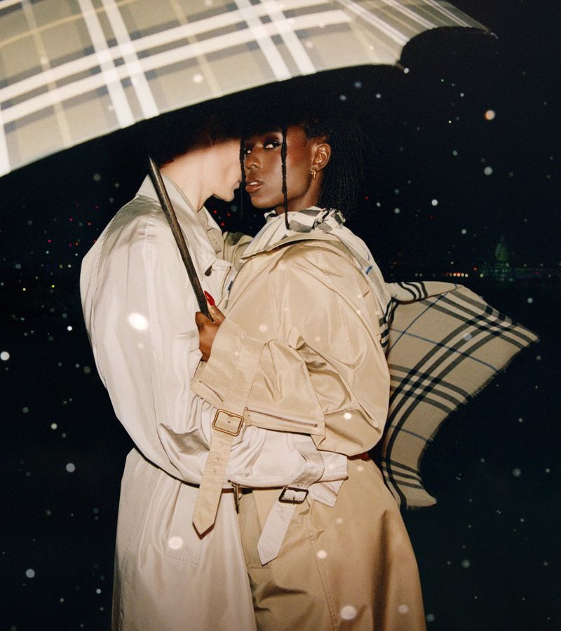 A couple poses beneath a large umbrella in the rain at night, both wearing tan trench coats in a fashion editorial setting.