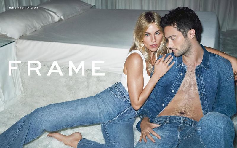 A blonde woman and a dark-haired man pose together on a white fluffy rug in an indoor setting, both wearing denim clothing. 