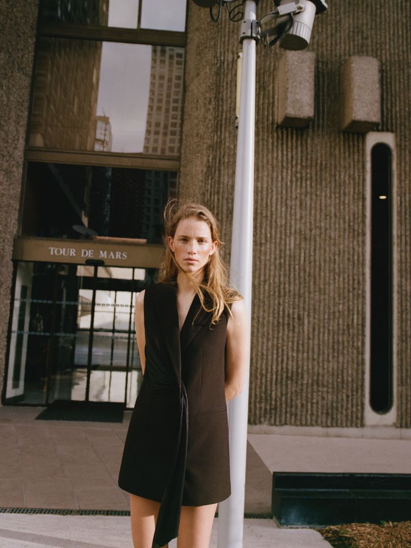 A fair-skinned young woman poses in front of a concrete building wearing a sleeveless black dress. A light pole stands nearby.