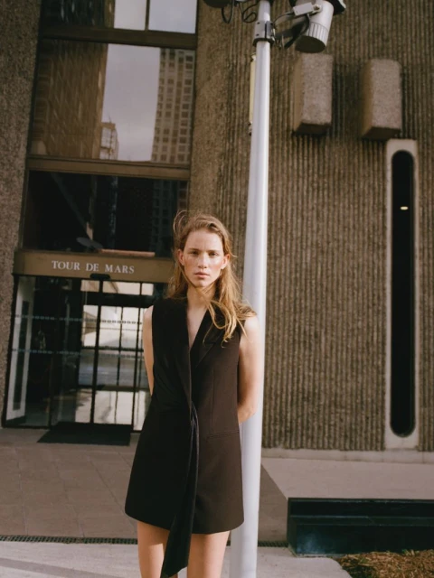 A fair-skinned young woman poses in front of a concrete building wearing a sleeveless black dress. A light pole stands nearby.