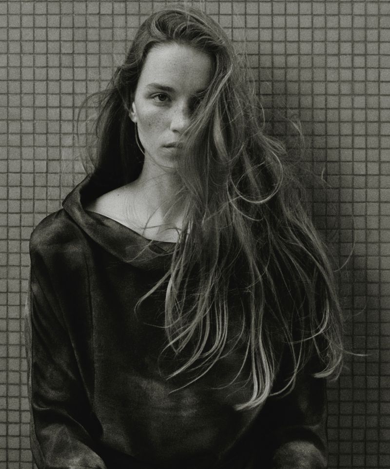 A portrait of a young adult woman with long hair wearing a dark sweater against a tiled wall in a close-up studio shot.