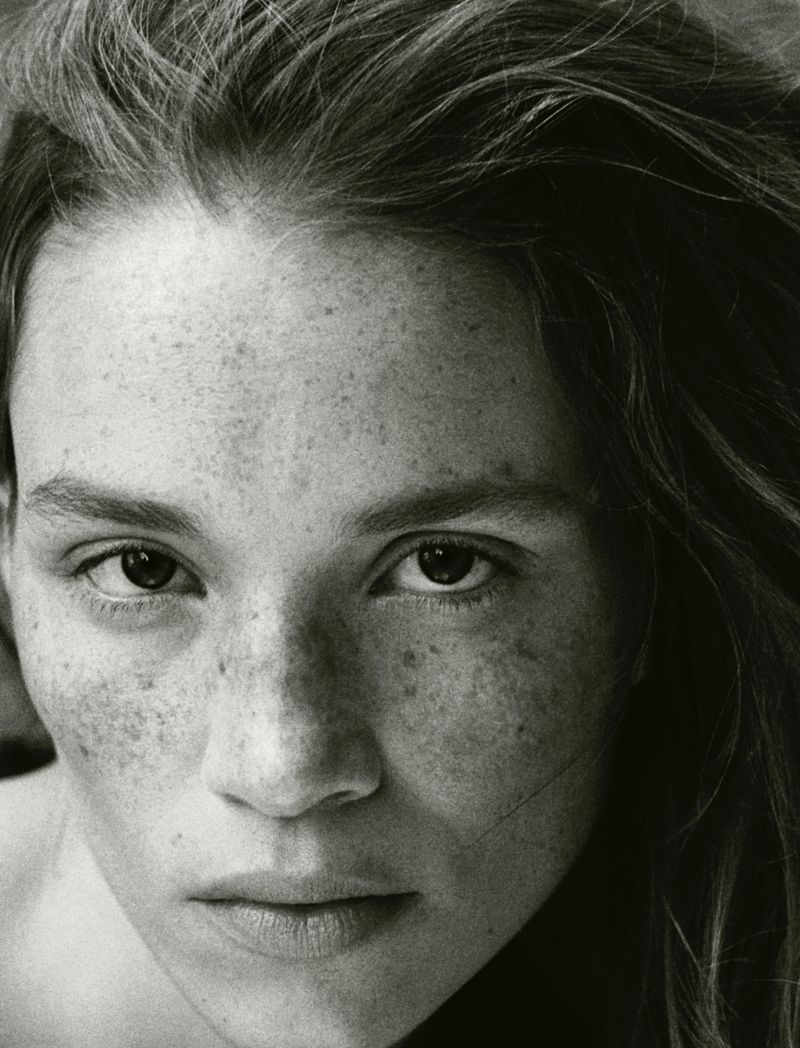 Close-up black and white portrait of a young adult female with freckles, gazing at the camera with a serious expression and loose wavy hair.