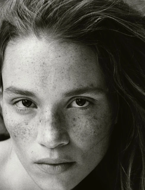 Close-up black and white portrait of a young adult female with freckles, gazing at the camera with a serious expression and loose wavy hair.