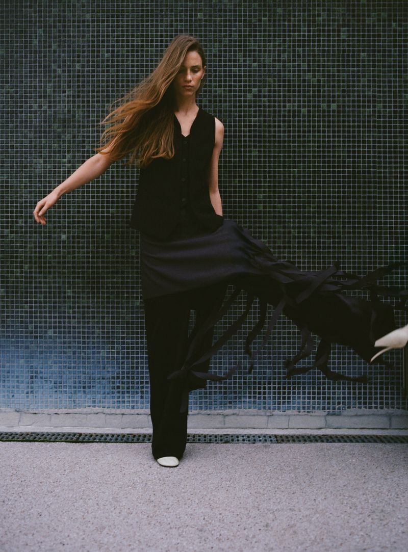 A stylish woman in dark, flowing clothing poses against a tiled wall with an outstretched arm in a fashion-forward shot.