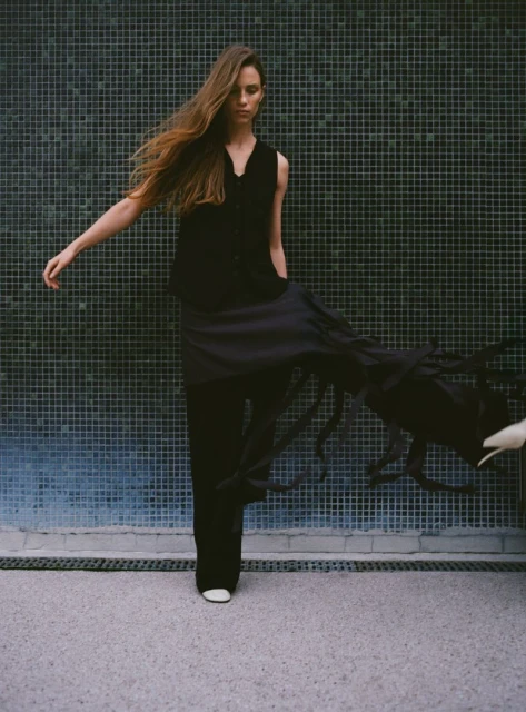 A stylish woman in dark, flowing clothing poses against a tiled wall with an outstretched arm in a fashion-forward shot.