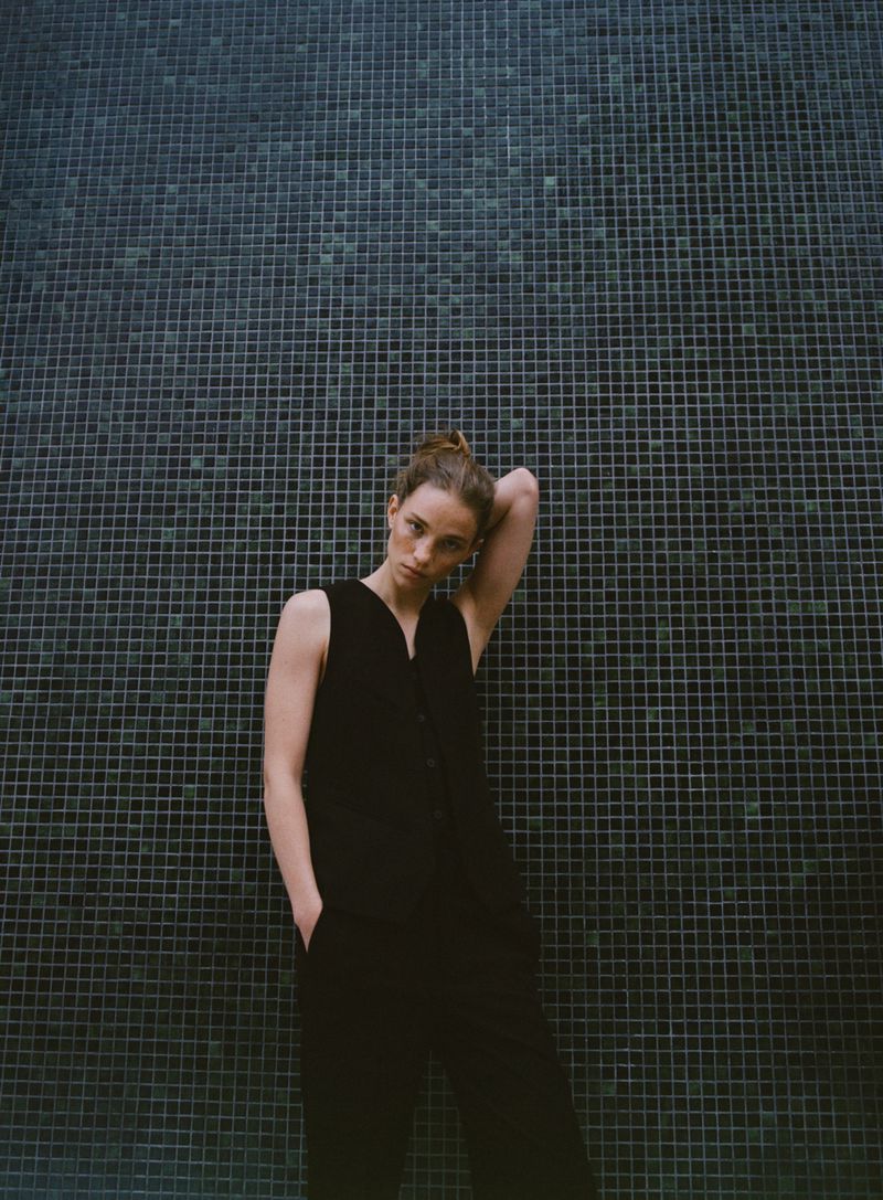 An adult female with hair in a bun poses in a sleeveless jumpsuit against a tiled wall in an eye-level, medium-shot composition.