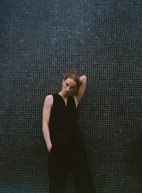 An adult female with hair in a bun poses in a sleeveless jumpsuit against a tiled wall in an eye-level, medium-shot composition.