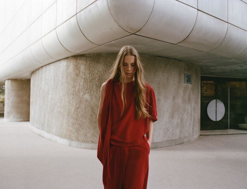 Woman with long blonde hair in a red outfit poses on a concrete building exterior. Fashion shot with natural light.