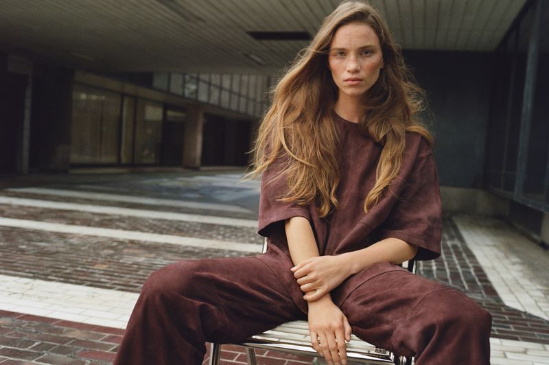 A female model with long brown hair, wearing a maroon t-shirt and pants, sits on a metal chair in an urban environment with muted tones.