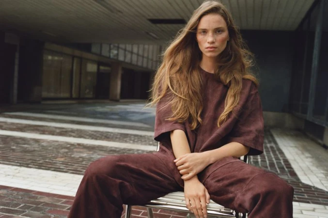 A female model with long brown hair, wearing a maroon t-shirt and pants, sits on a metal chair in an urban environment with muted tones.