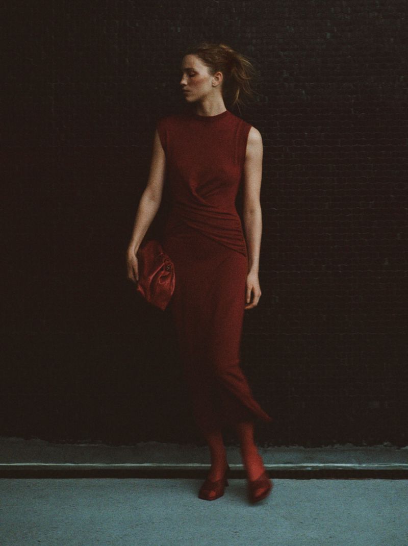 Elegant woman in a maroon dress and matching accessories poses against a dark textured wall, showcasing fashion and style.