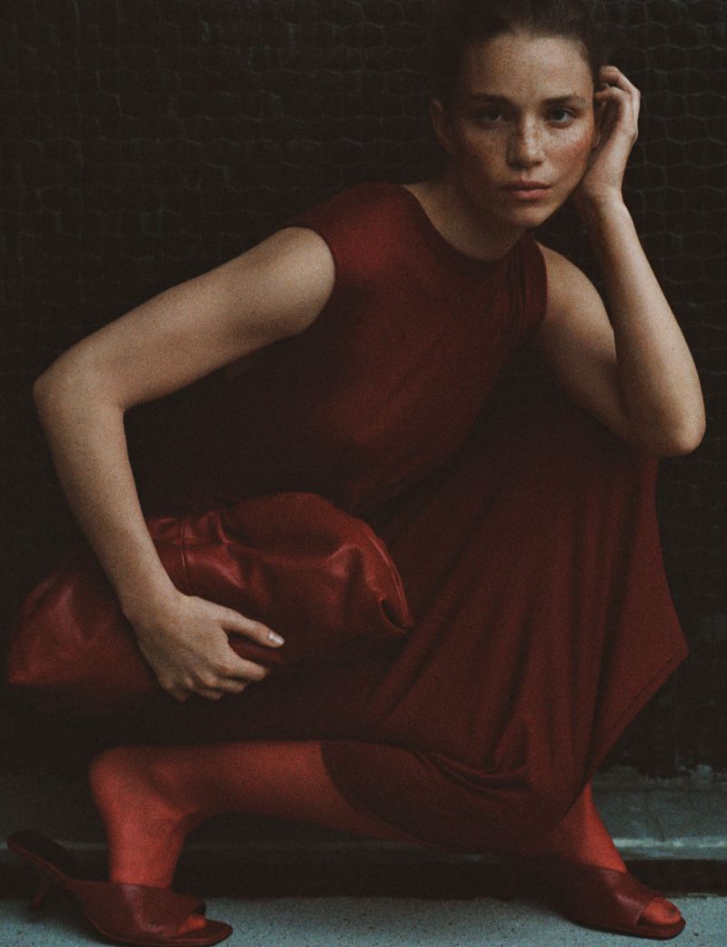 Portrait of a model in a red dress and shoes, crouching against a dark wall with a matching red bag. Fashion shot, medium angle.