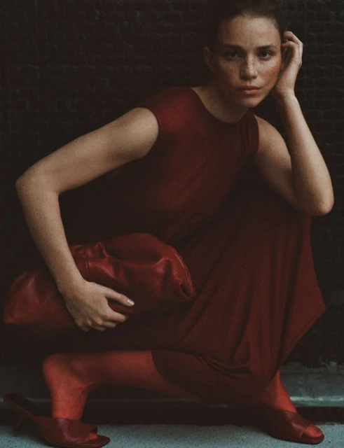 Portrait of a model in a red dress and shoes, crouching against a dark wall with a matching red bag. Fashion shot, medium angle.