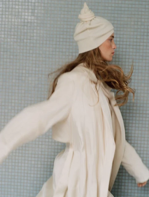 A light-skinned woman with flowing light-brown hair, wearing a white outfit and head covering, strikes a pose against a pale blue tiled wall.