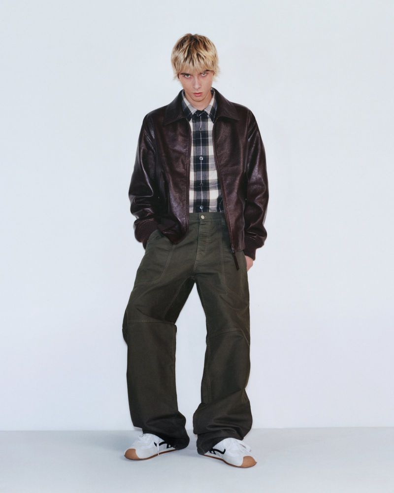 Male fashion model in brown leather jacket, plaid shirt, and olive trousers posing in a full shot against a white backdrop.