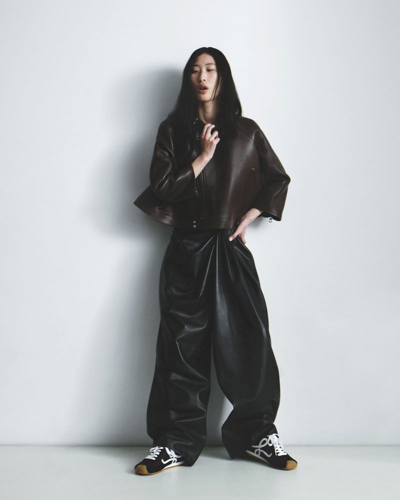 Model wearing a dark brown leather jacket and matching pants with black sneakers against a plain white studio background.