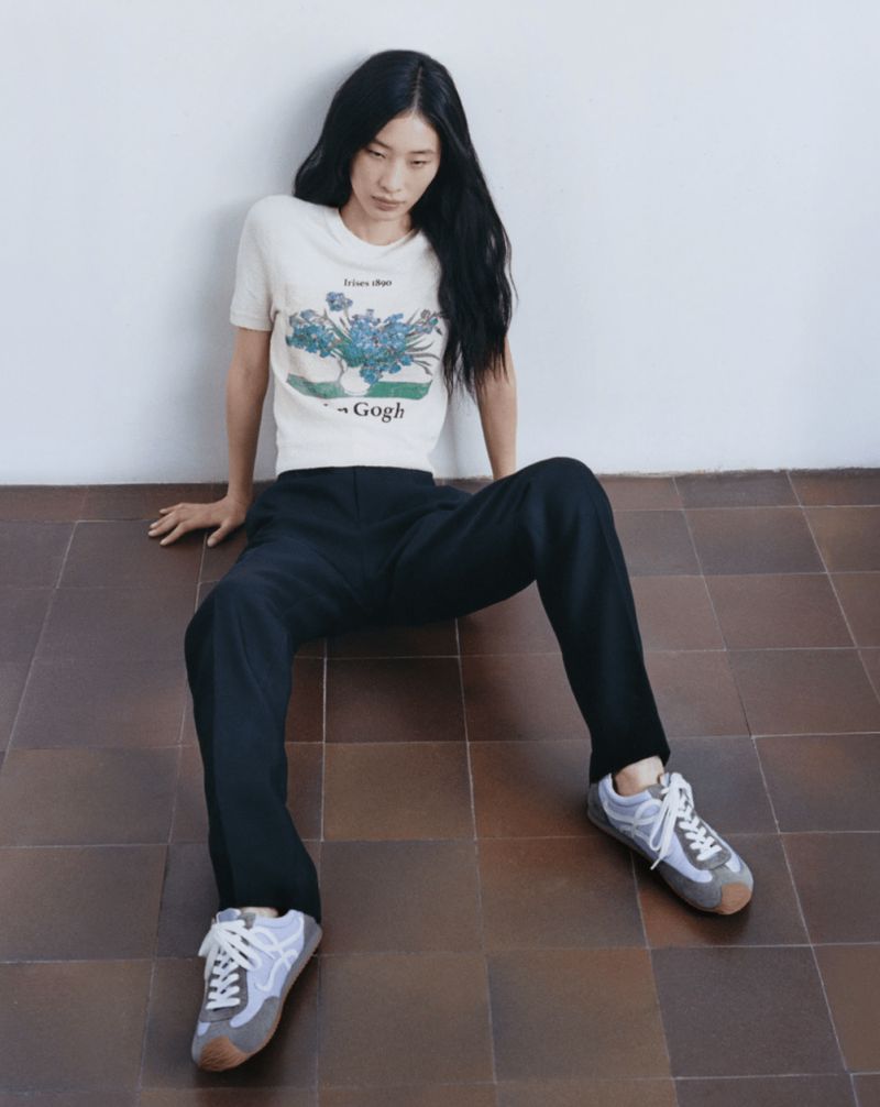 Asian woman in a casual studio shot wearing a graphic tee, black pants, and sneakers, sitting on a brown tiled floor against a white wall.