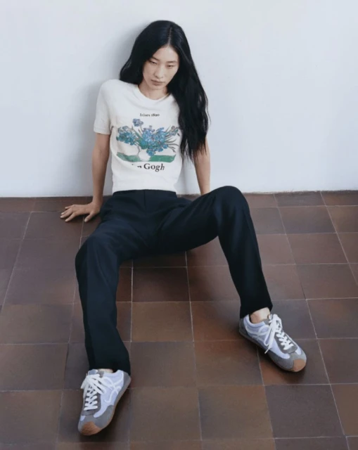 Asian woman in a casual studio shot wearing a graphic tee, black pants, and sneakers, sitting on a brown tiled floor against a white wall.