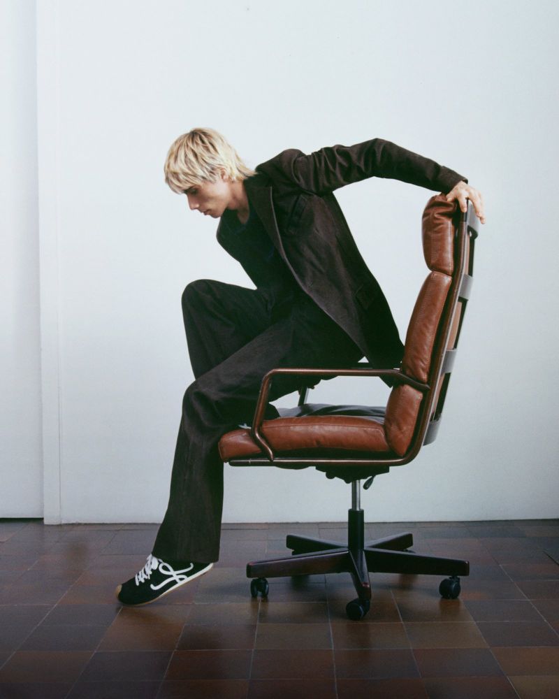 Male model with blond hair in a brown suit posing on a brown leather office chair against a white background in a studio.