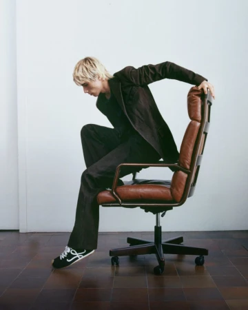 Male model with blond hair in a brown suit posing on a brown leather office chair against a white background in a studio.