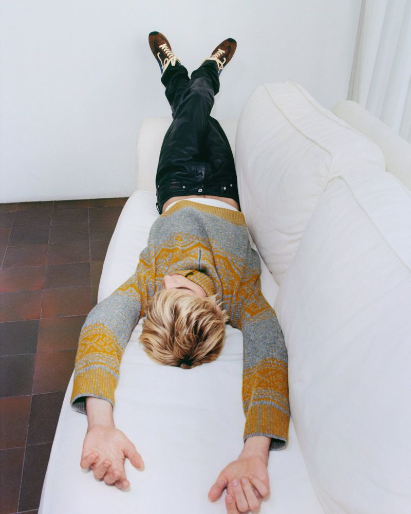Young man lying upside down on a white couch in a gray and yellow patterned sweater and black pants with brown shoes indoors.