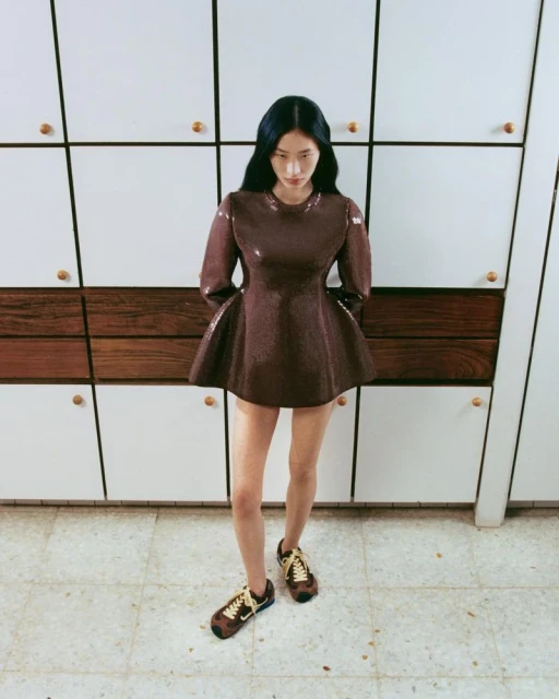 An Asian woman in a brown dress and sneakers stands in a white room with white and wood cabinets, looking down in a fashion editorial shot.