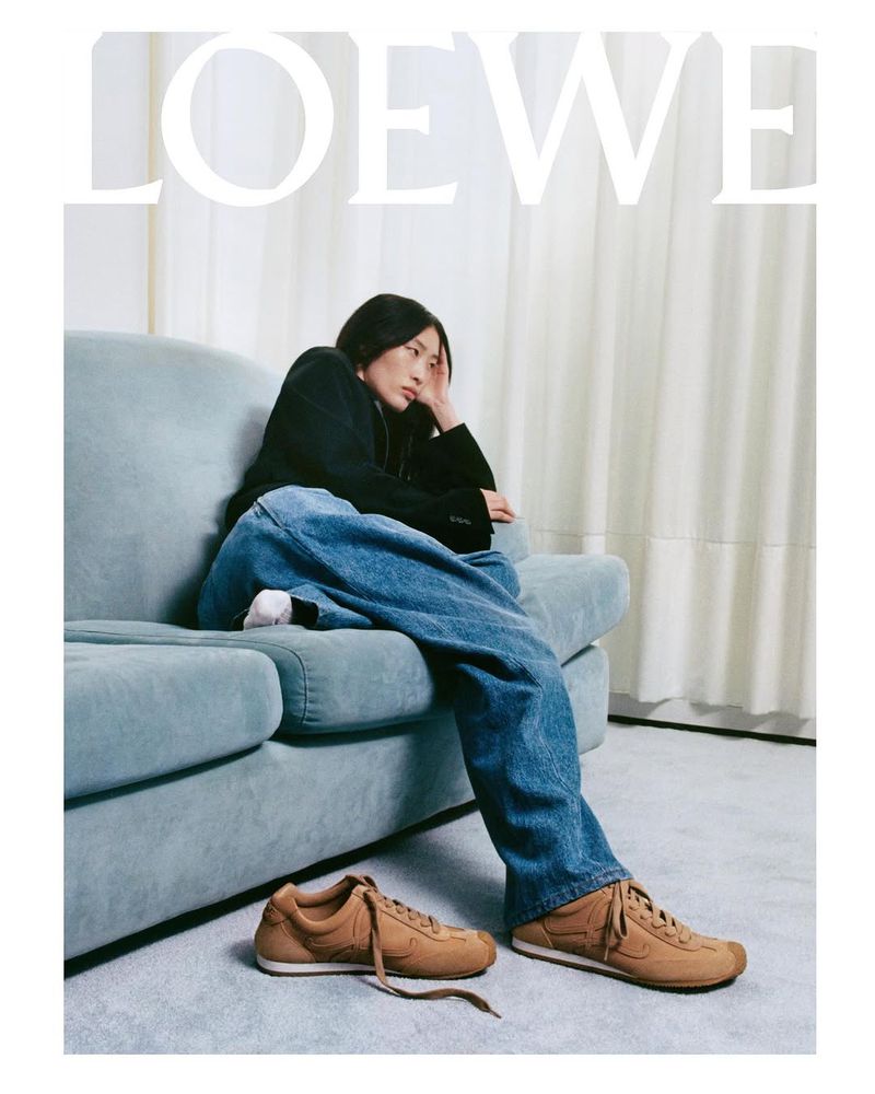 Woman sits on a light-blue sofa in jeans, with brown sneakers on the floor in an indoor setting; brand logo 'LOEWE' is visible.