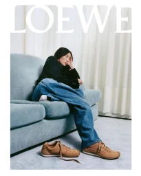 Model seated on a light blue sofa wearing a dark jacket and wide leg blue denim jeans with tan sneakers
